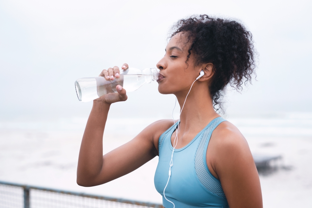 Woman, drinking water and exercise in outdoor for hydration, music and runner or workout break. Female person, profile and relax on promenade, cloudy sky and bottle of mineral liquid for nutrition